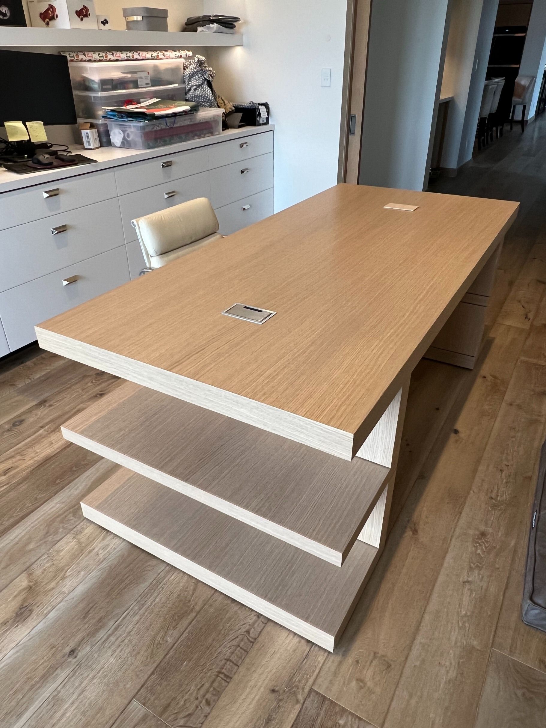 A light-wood desk with built-in side shelves and integrated power outlets, located in a home office with white cabinetry.