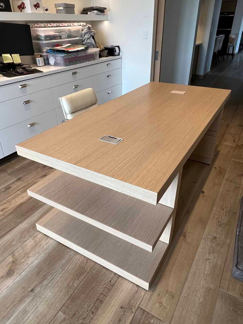 A light-wood desk with integrated open shelving at one end, positioned in a modern office or study space.