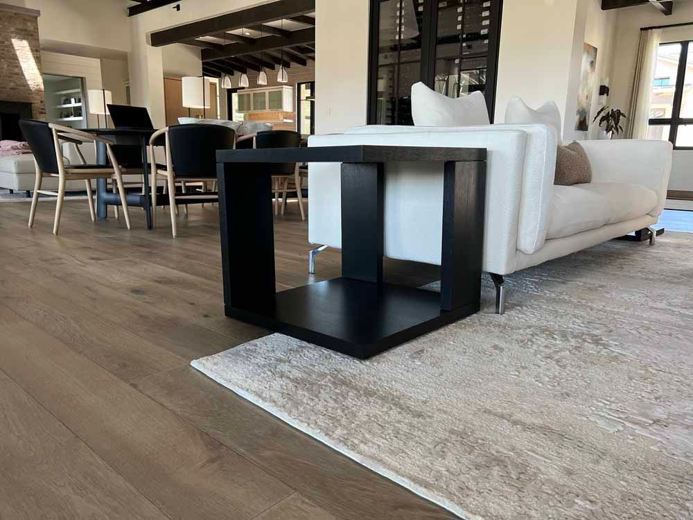 A modern black square end table sits on a beige rug next to a white sofa in a bright, open-plan living and dining room.