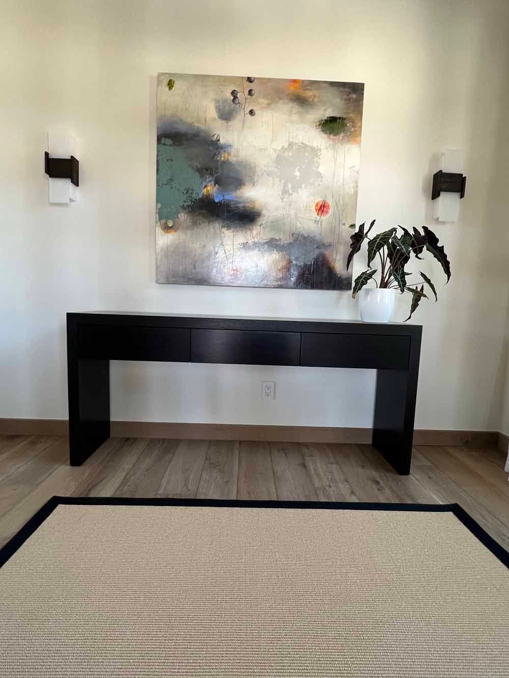 A long black console table sits against a wall beneath an abstract painting, with a potted plant and two wall sconces.