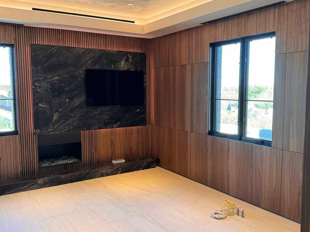 A room with wood-paneled walls, a black marble accent wall featuring a television and fireplace, and a large window.