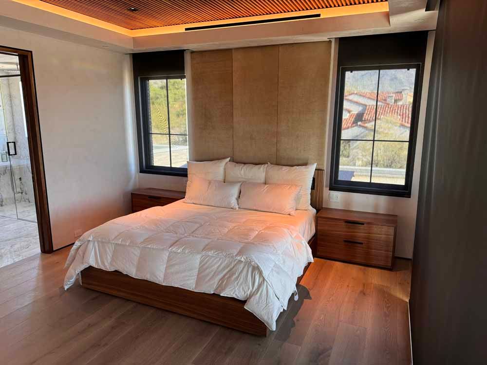 A modern bedroom features a bed with a white comforter, a tan upholstered headboard, two wooden nightstands, and windows.