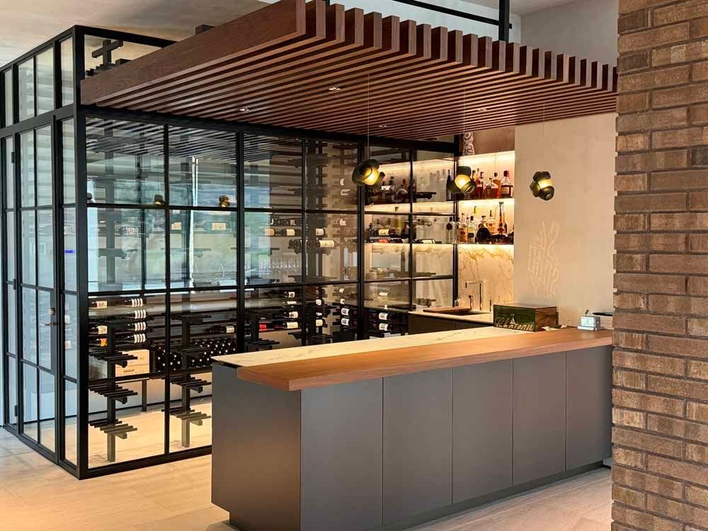 Modern home bar with a charcoal gray counter, a slatted wood ceiling, glass-walled wine storage, and a brick accent wall.
