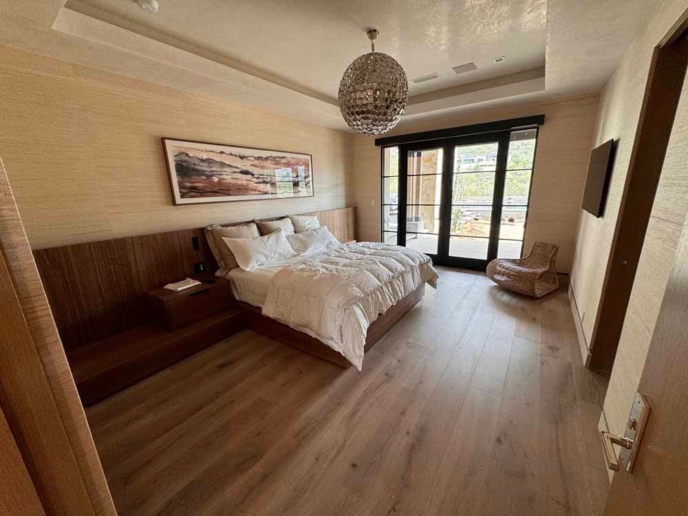 A modern, neutral-toned bedroom with a large bed, wooden headboard, chandelier, and glass doors leading to a balcony.