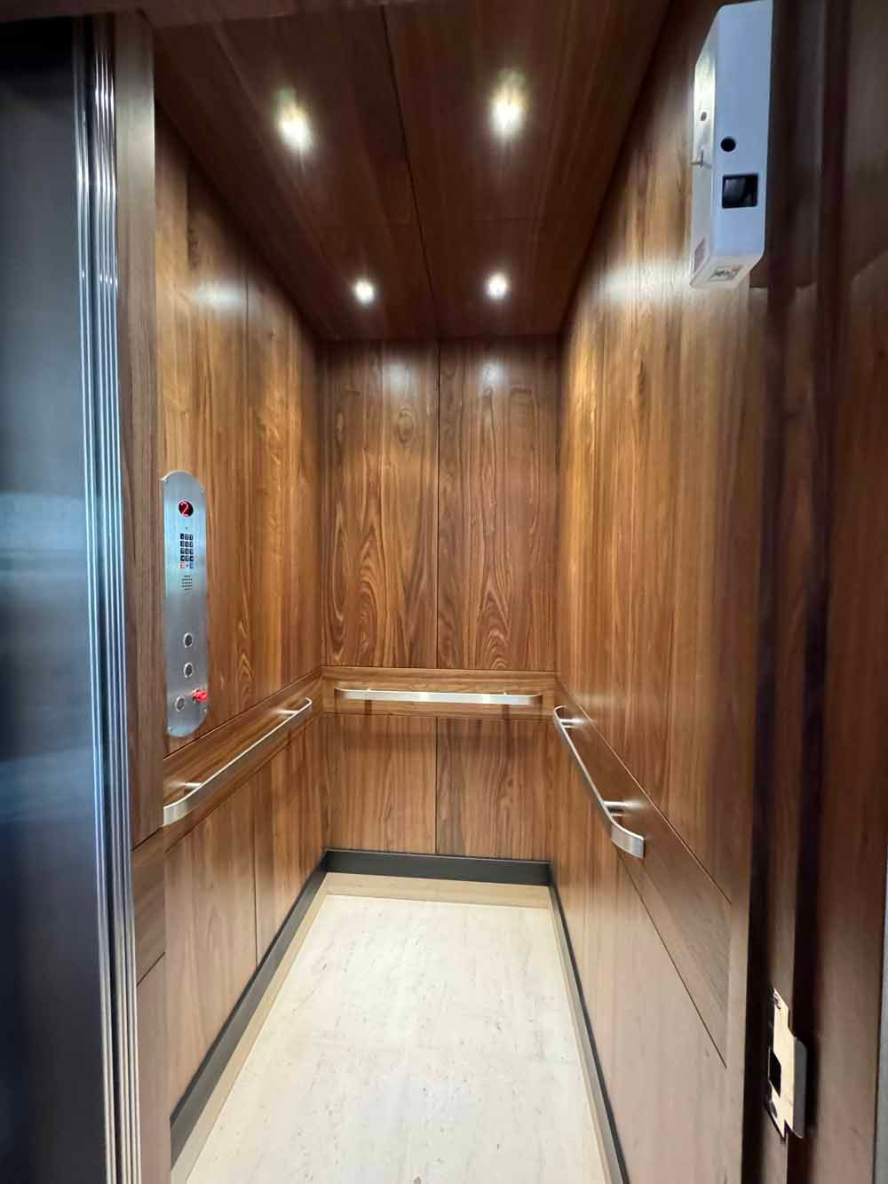 Interior of an elevator with wood-paneled walls, warm recessed ceiling lights, stainless steel accents, and light flooring.