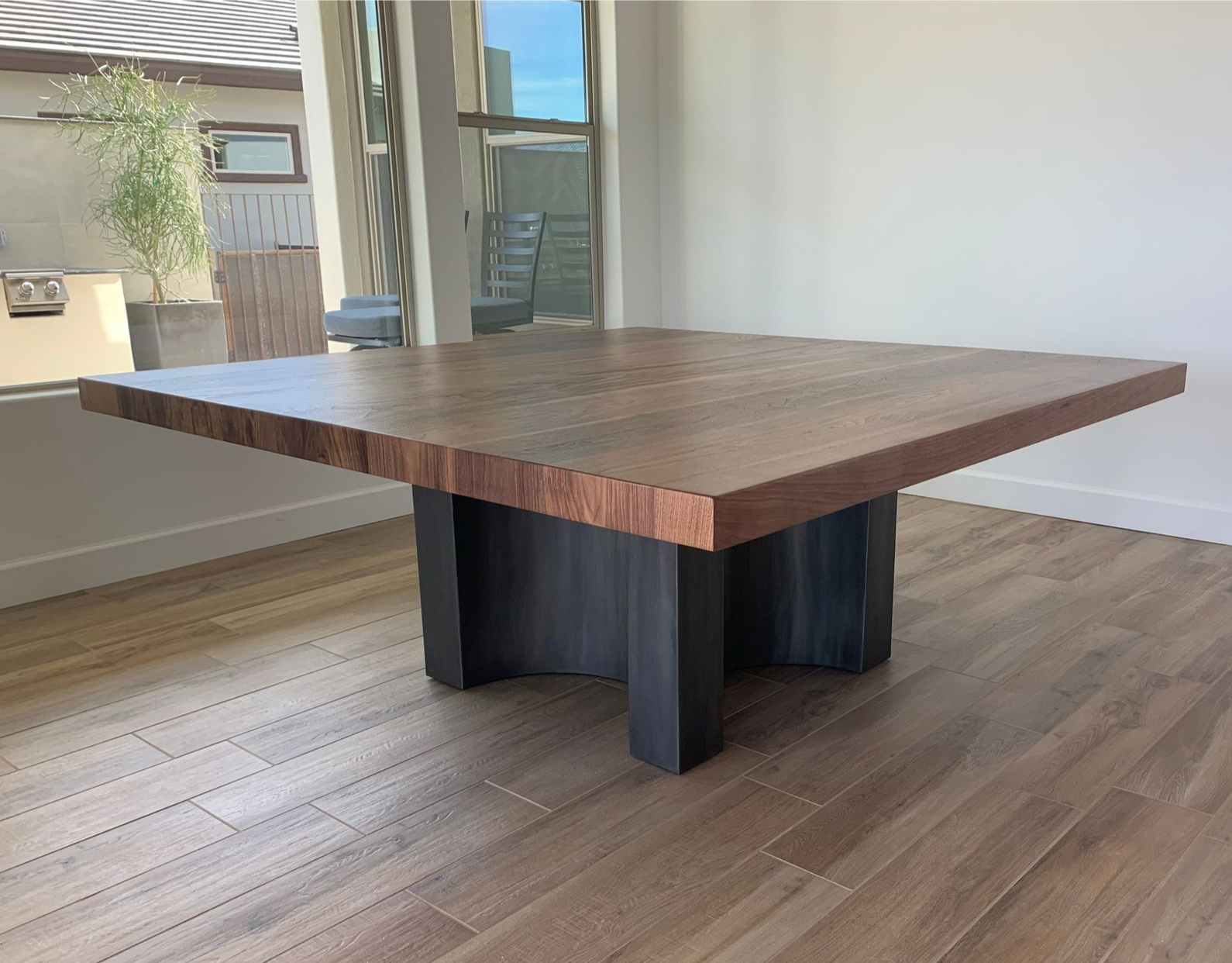 A square wooden coffee table with a thick grain top and a dark, heavy, cross-shaped metal base in a bright room.