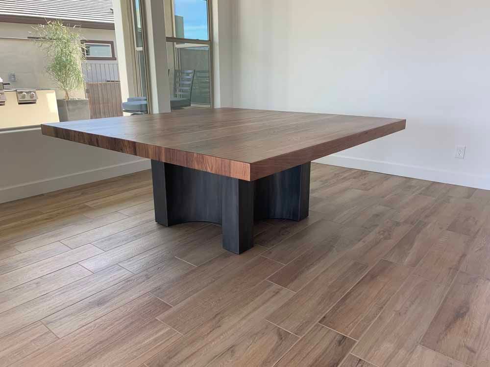 A large square wooden table with a thick dark brown top and a black industrial-style base stands on a wood-look floor.