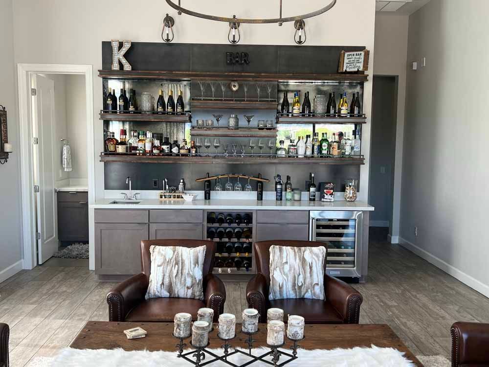 A home bar area featuring wood cabinets, floating shelves with glass decor, wine storage, and two brown leather chairs.