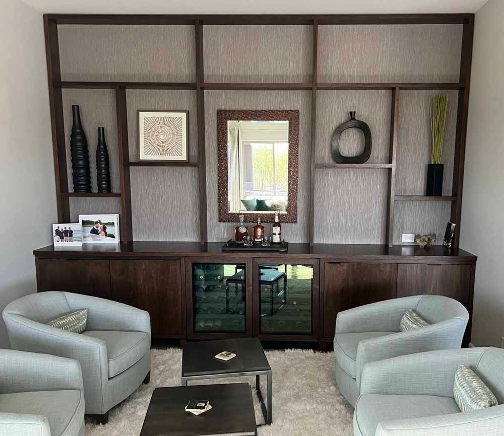 Four gray armchairs surround two small black tables in front of a floor-to-ceiling dark wood shelving unit with a bar area.