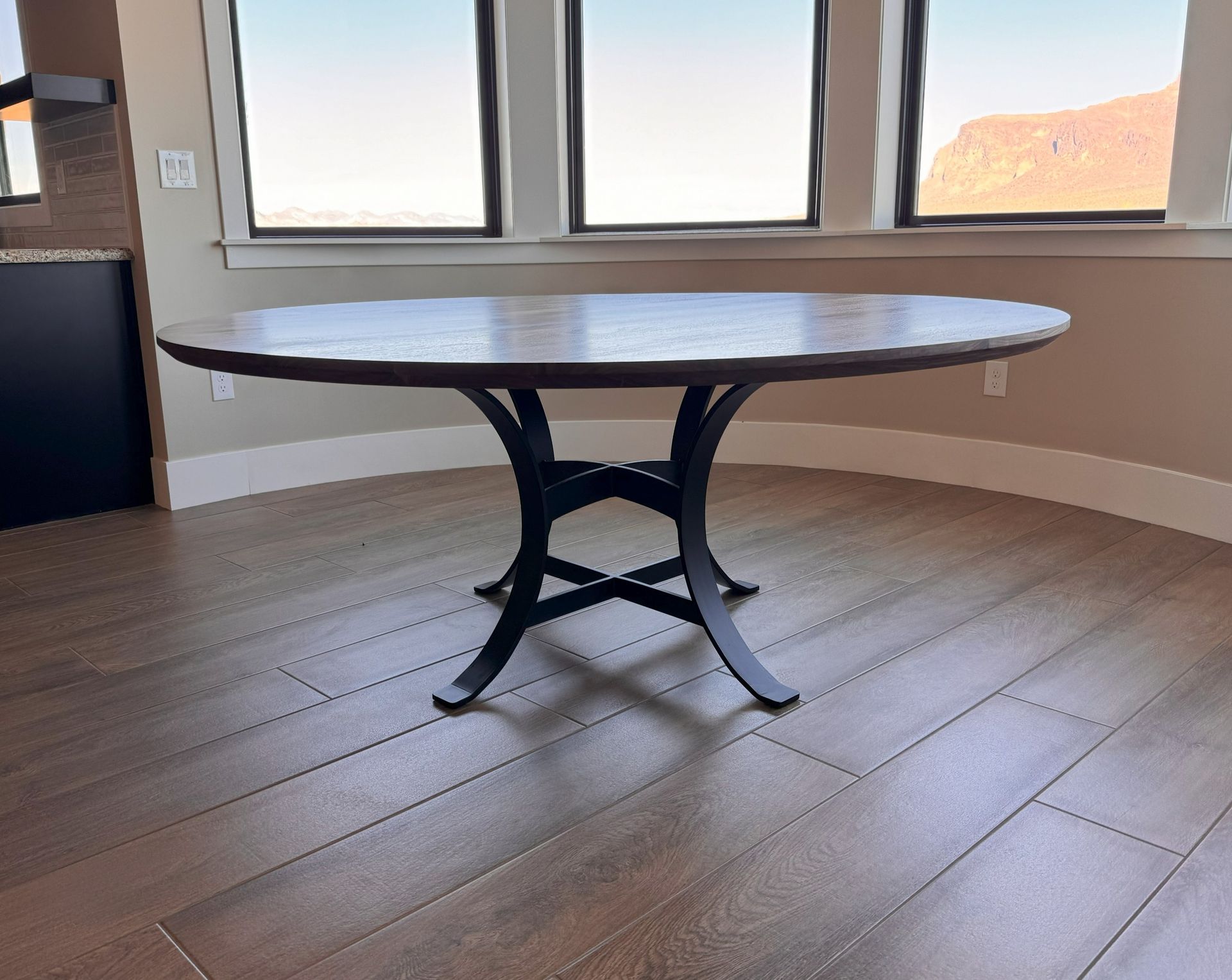 A round wooden dining table with a black, curved metal pedestal base stands in a room with hardwood floors and windows.
