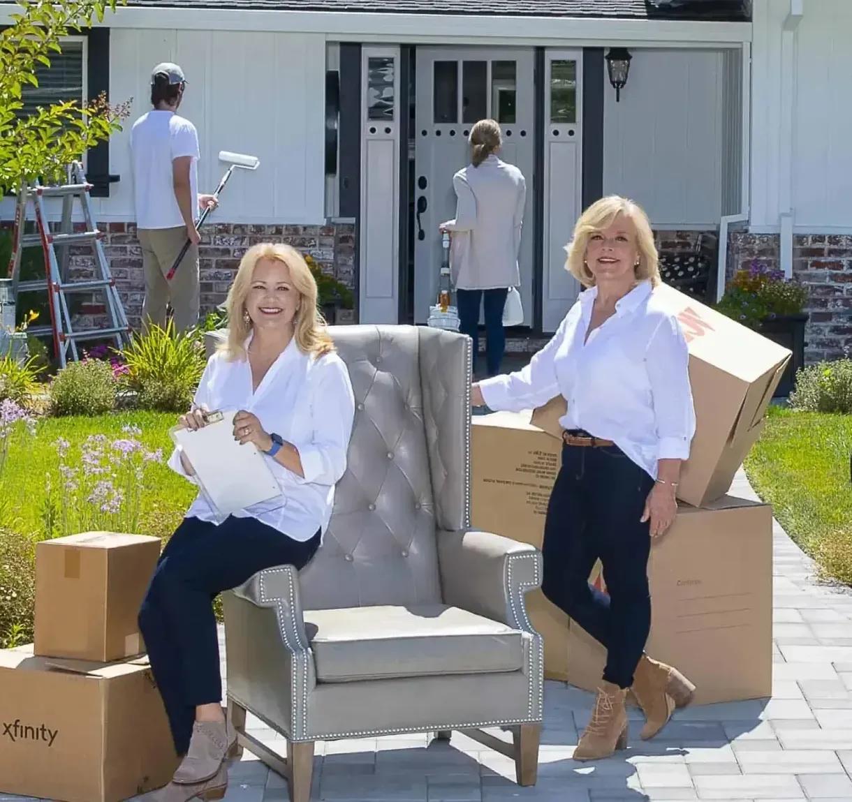 Two women with boxes and furniture in front of a house; workers are painting and standing by the front door.
