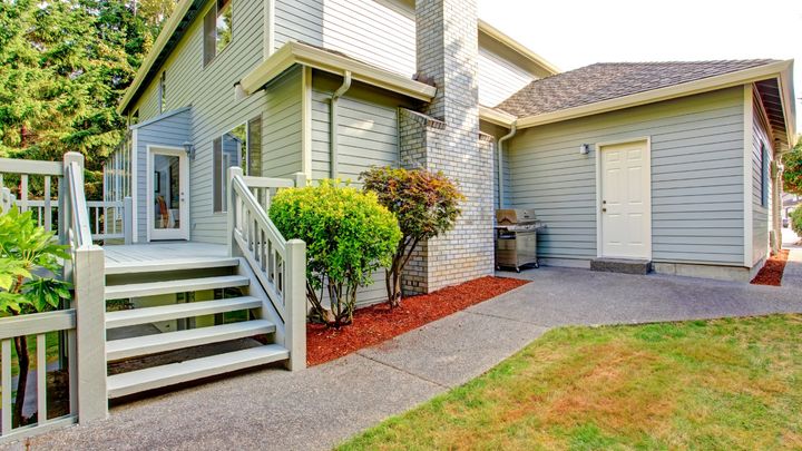 A small white wooden deck attached to the back of a house.  There is wooden stairs leading down to a concrete walkway.