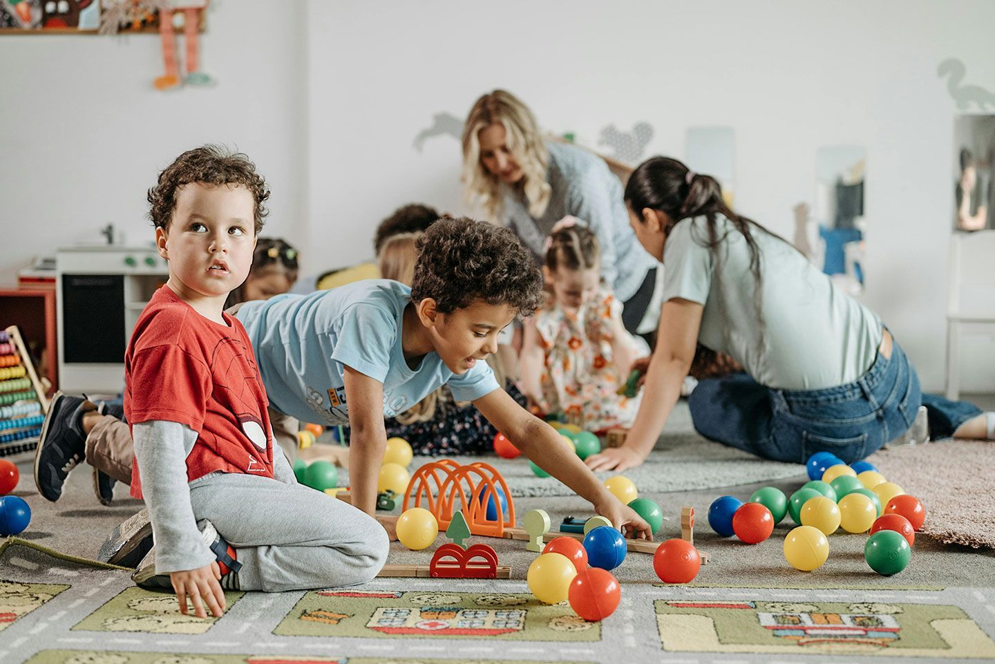 Children playing with colorful toys on a classroom floor while adults supervise in the background