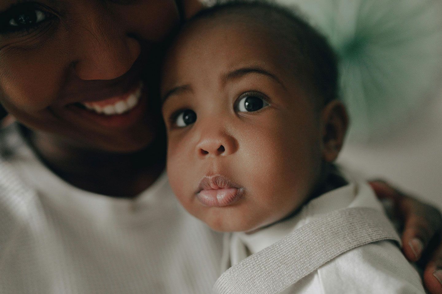 Close-up of a baby in a white outfit being held, with an adult smiling in the background.
