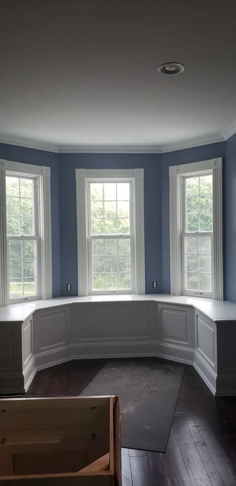 Bay window nook with white built-in bench, blue walls, dark wood floors, and three windows