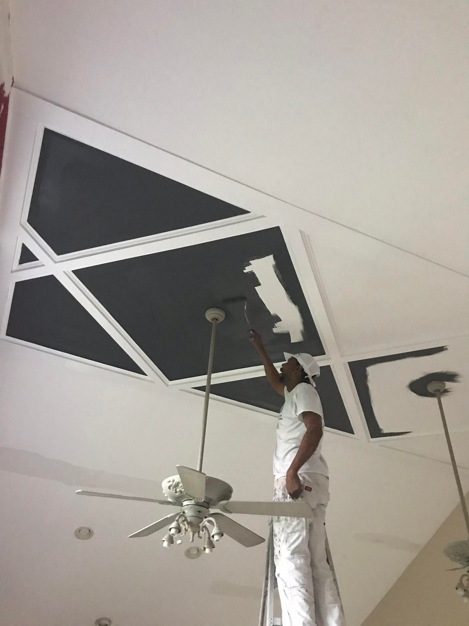 Person in white clothing painting a black geometric ceiling design beside a ceiling fan.