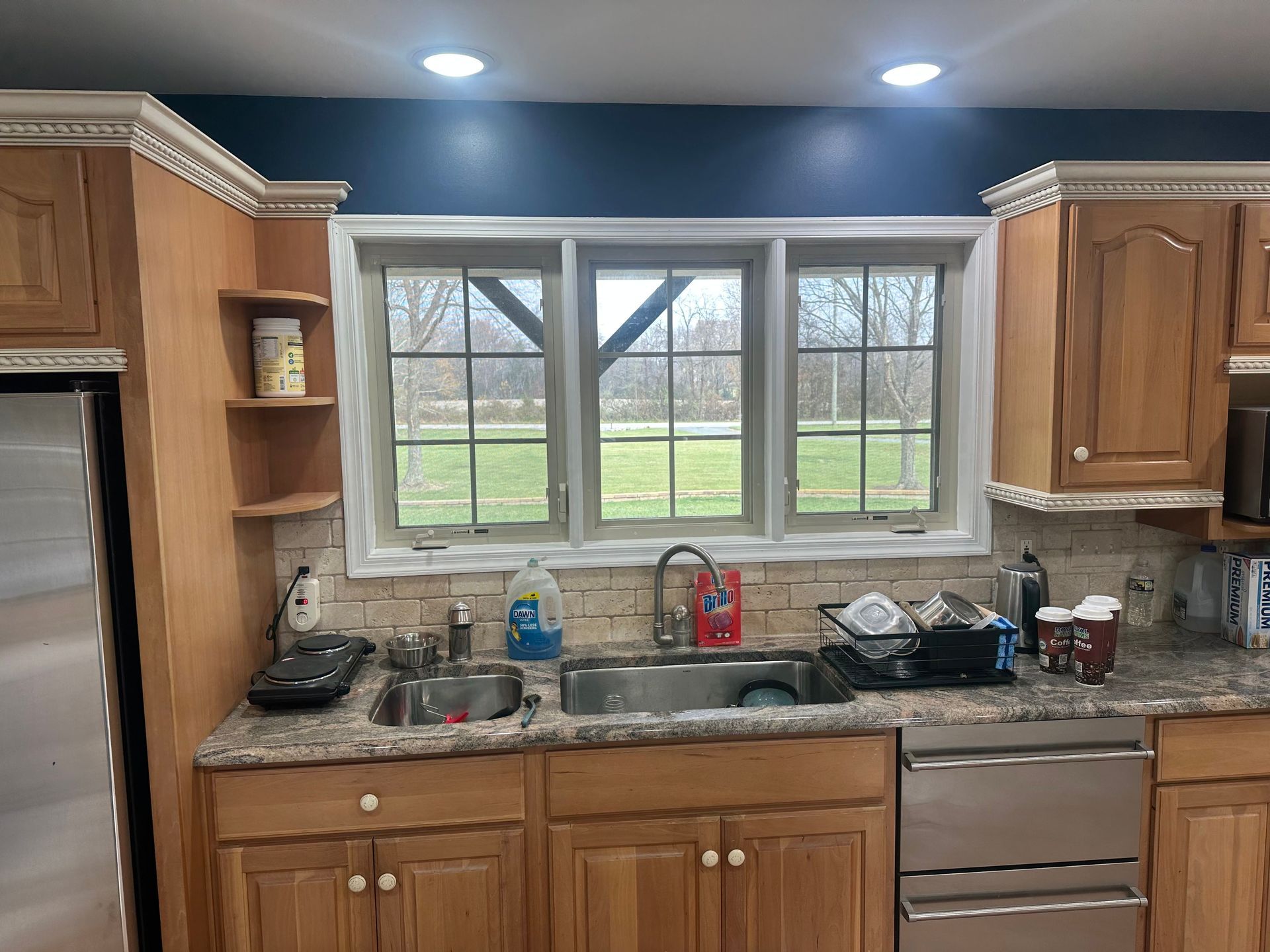 Kitchen sink under window with granite countertop, wood cabinets, and stainless dishwasher under blue ceiling lights