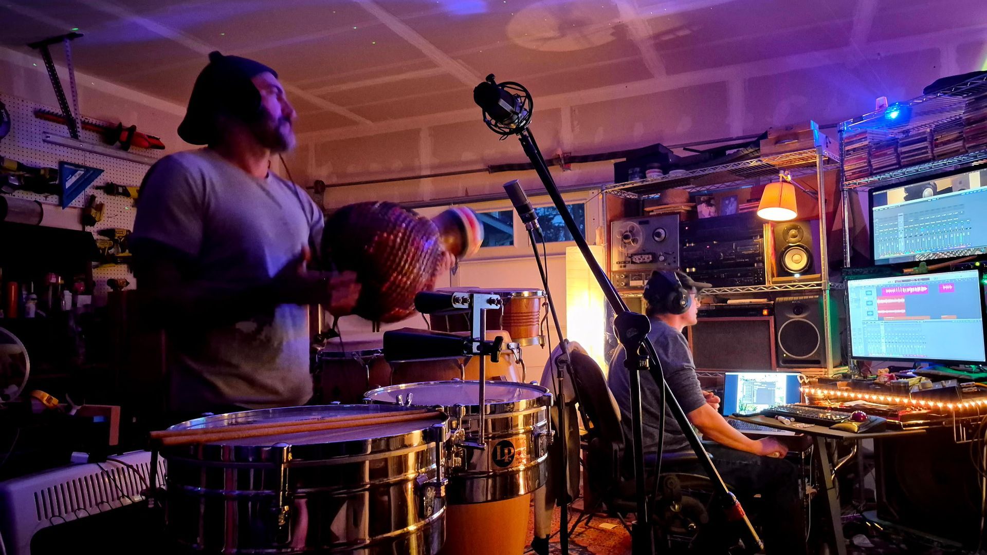 Drummer performing in a dim, colorful studio with microphones and computer monitors nearby