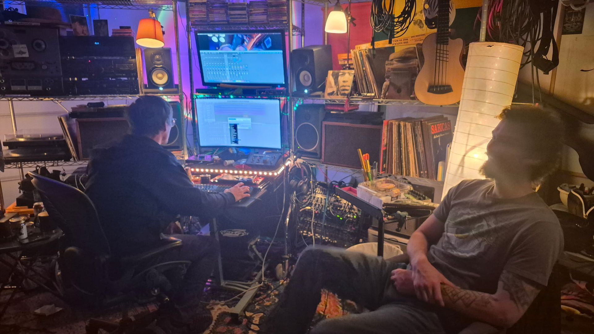 Two people in a dimly lit, cluttered music studio, one at a computer and one relaxing in a chair.