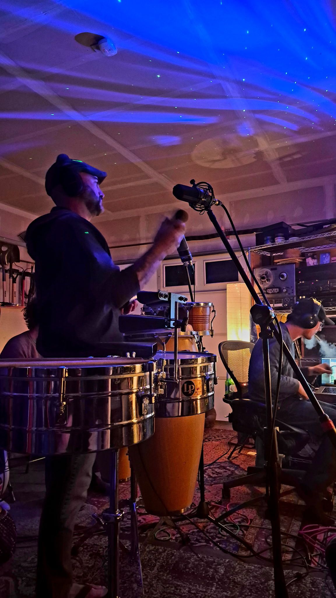 Musician playing conga drums onstage under blue-purple lights, with a microphone stand nearby