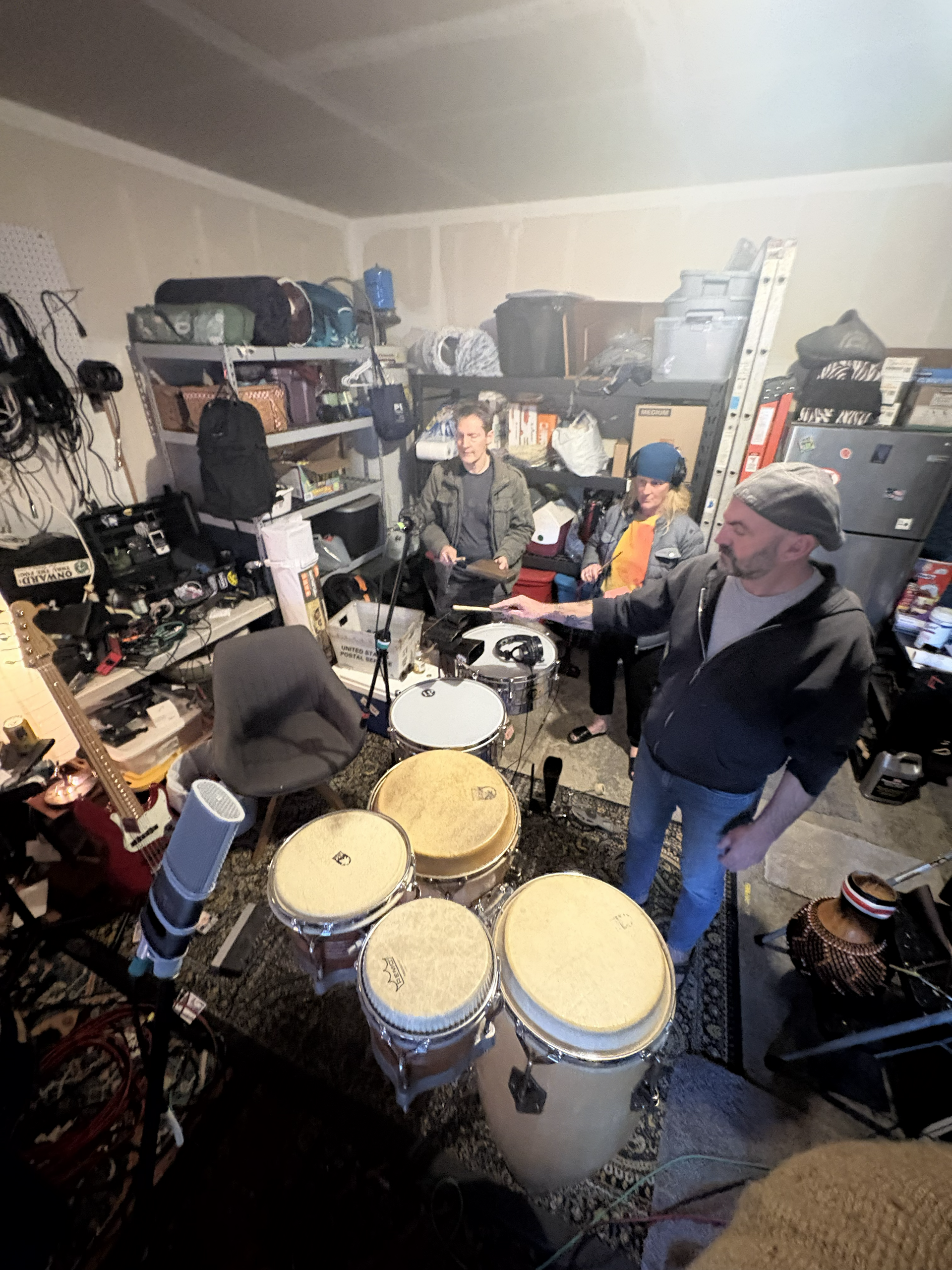 Cluttered garage rehearsal space with drums, shelves, and two people standing among musical equipment