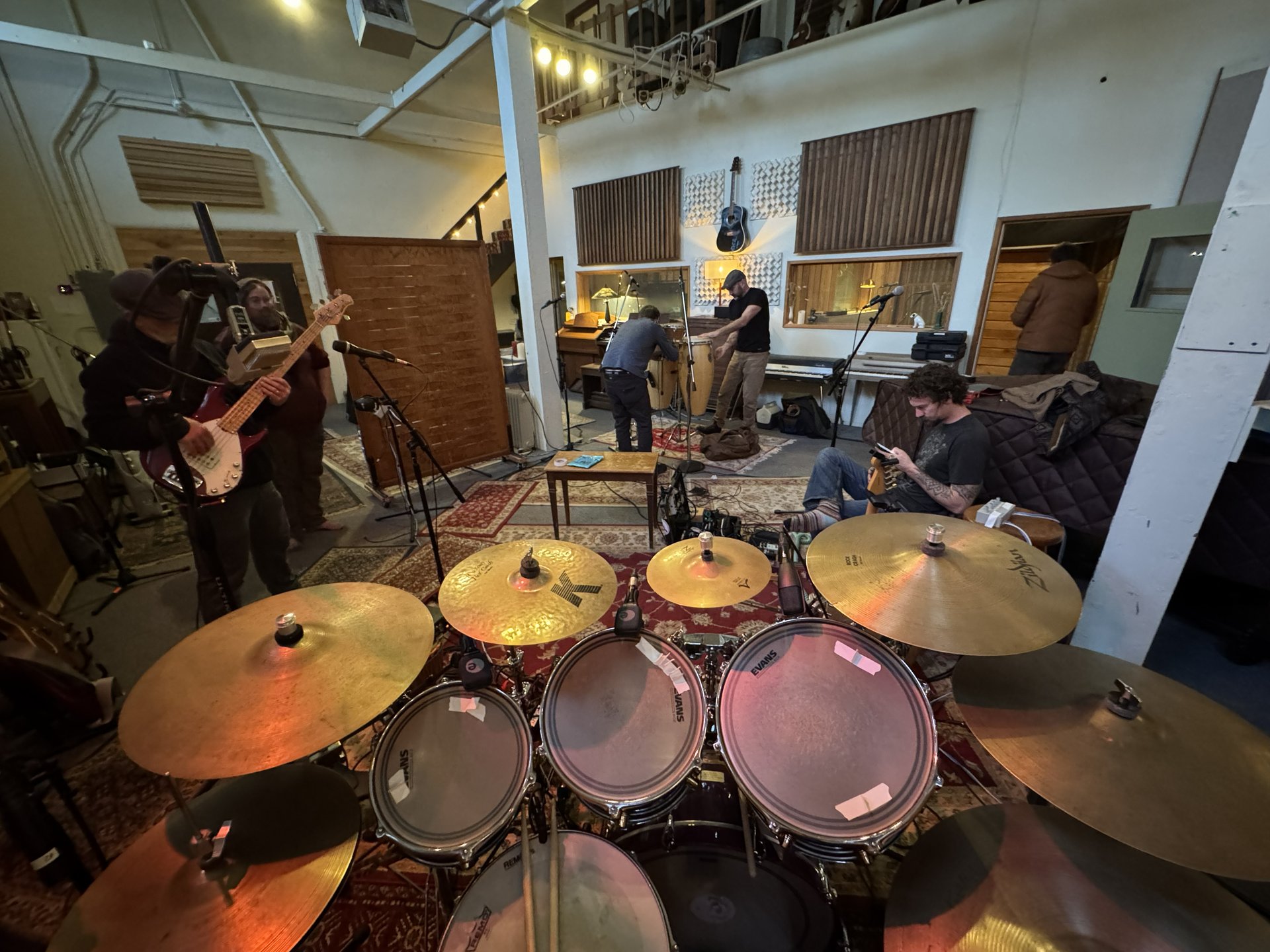 Band recording in a studio, viewed from behind a drum kit with cymbals in the foreground.