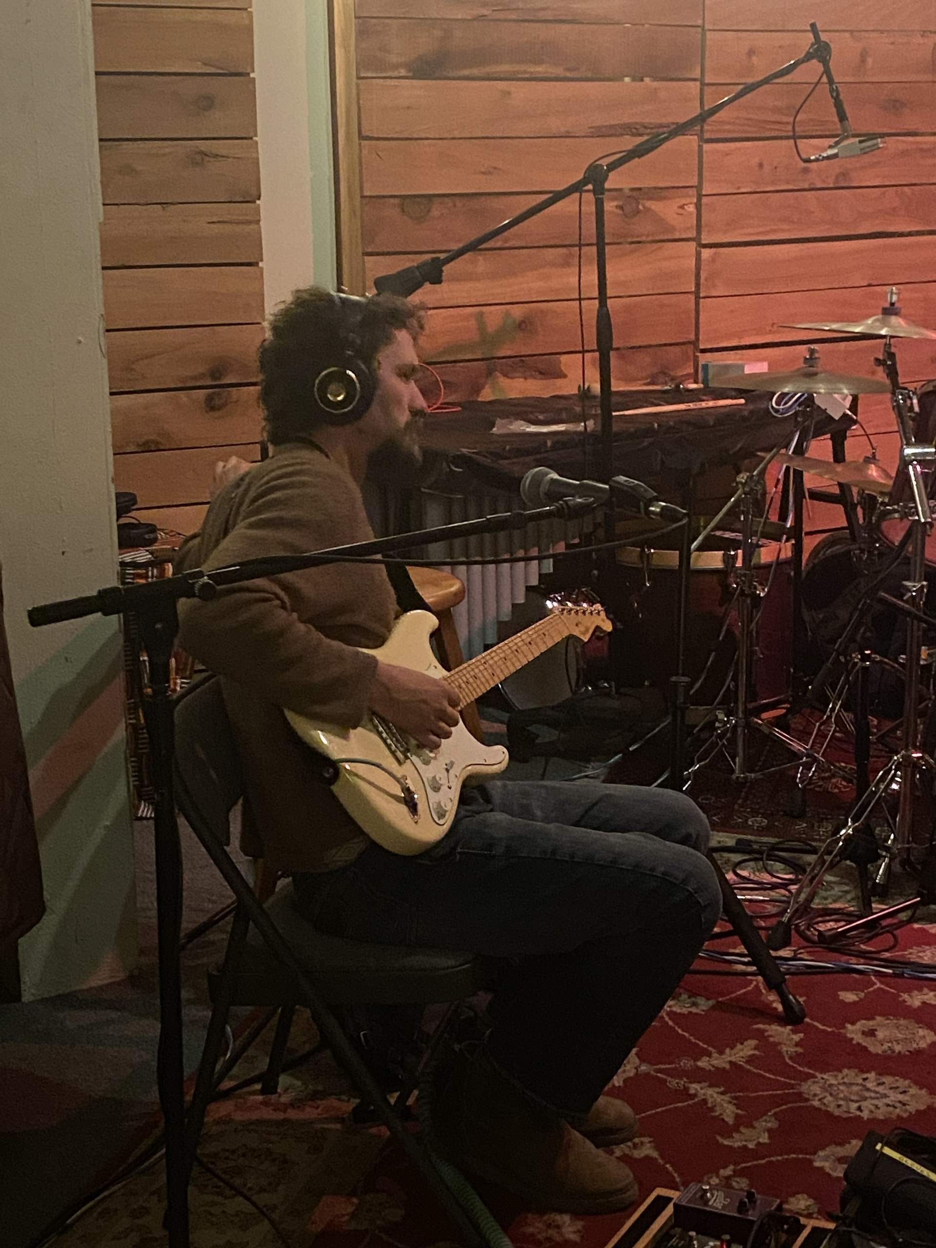 Musician seated in a studio, playing electric guitar with headphones near a drum kit and microphone stands