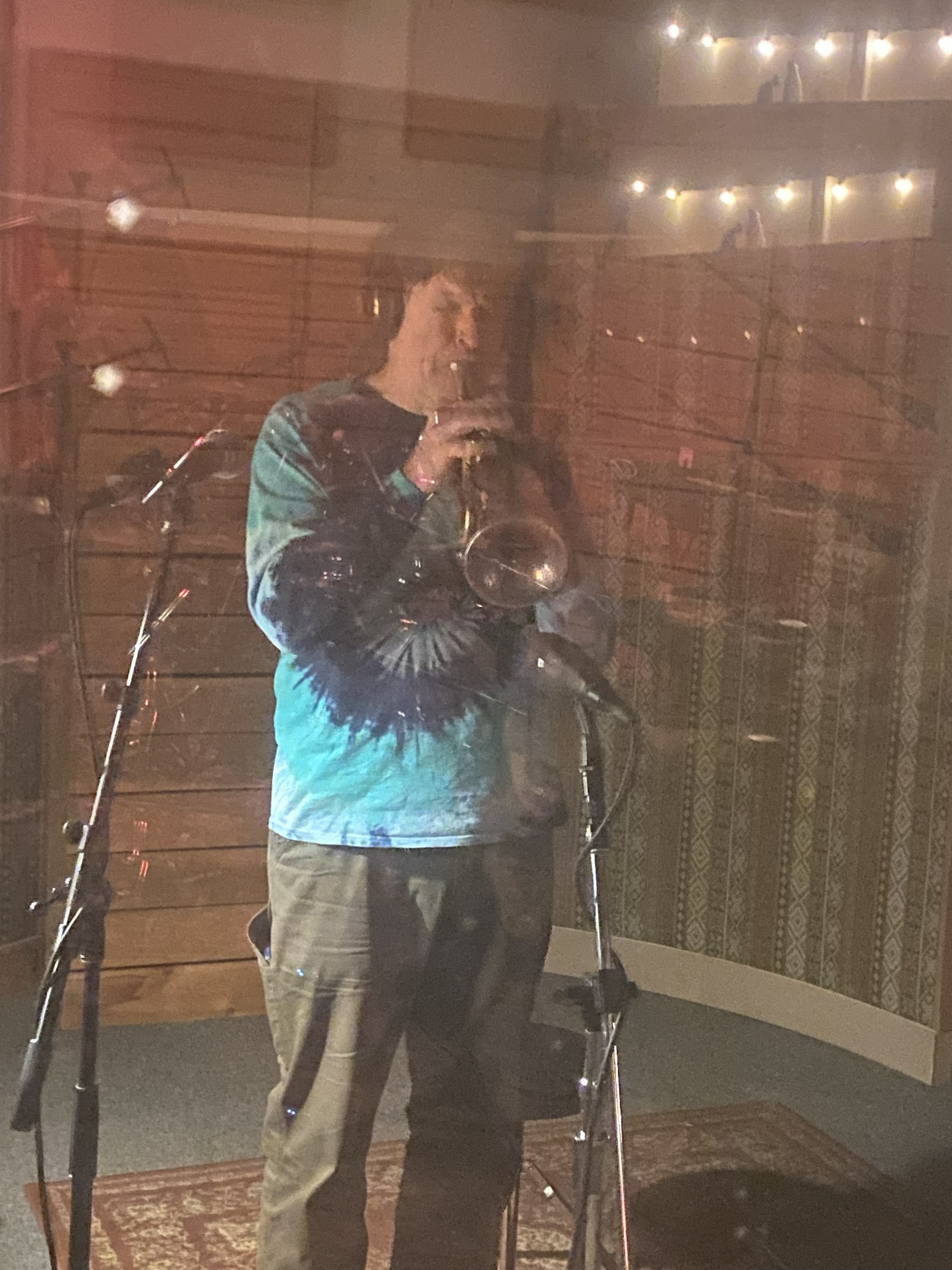 Person playing trumpet in a recording booth, wearing a blue tie-dye shirt and headphones.