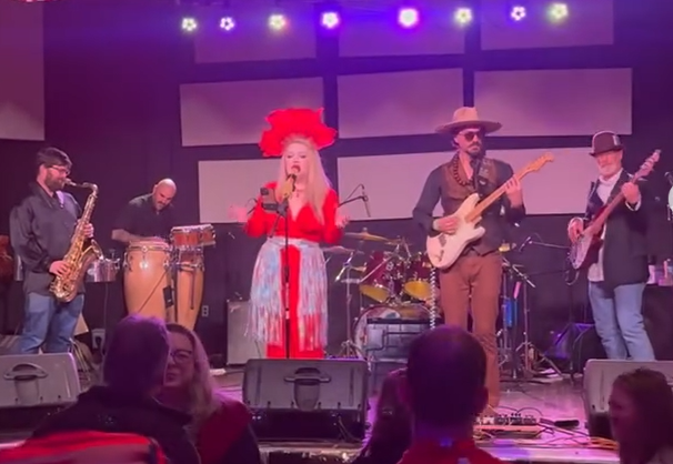 Live funk band performing on stage with purple lights; singer in red dress and hat, musicians playing sax, trumpet, drums, congas, bass, and guitar.