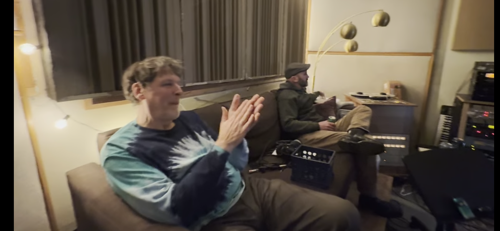Two people sitting in a living room, one clapping while the other works on a laptop.