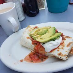 Breakfast wrap topped with avocado and sour cream on a white plate beside a mug