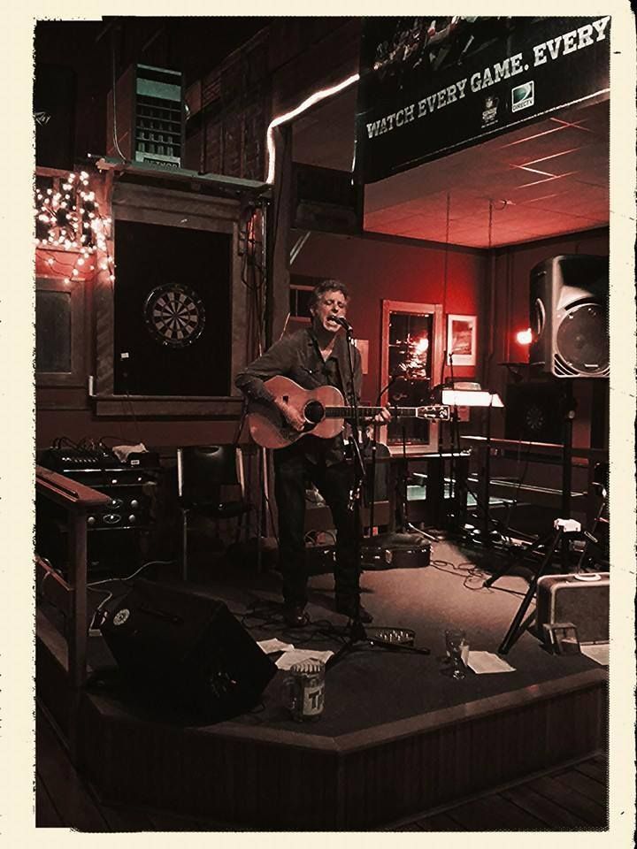 Solo musician performing with guitar on a dim red-lit stage in a small bar, with a dartboard nearby