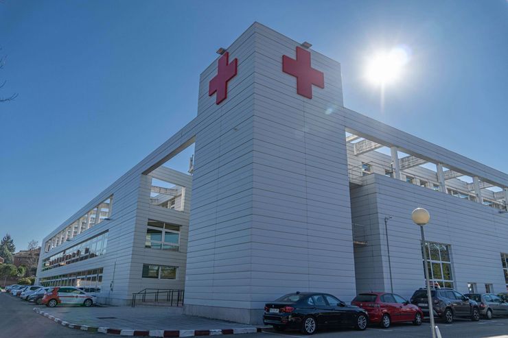 Edificio hospitalario moderno con símbolos de la cruz roja, coches aparcados y luz solar brillante.