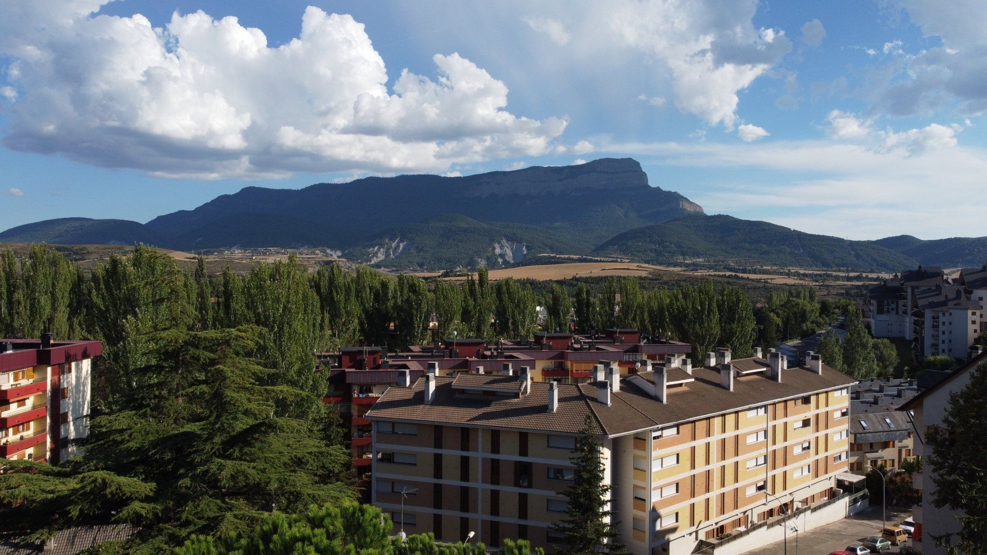 Horizonte montañoso sobre un pueblo con árboles, edificios de apartamentos y un cielo azul nublado.