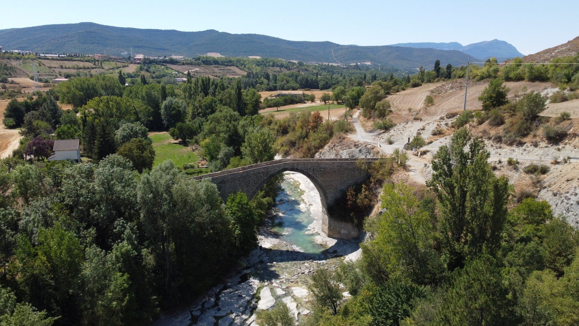 Puente de piedra sobre un río en un valle seco con árboles y colinas a lo lejos bajo un cielo despejado.