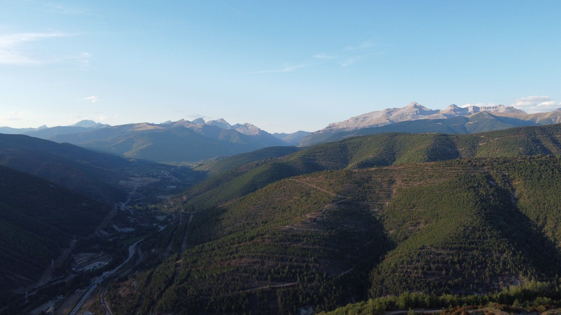 Valle de montaña con laderas boscosas y picos nevados a lo lejos bajo un cielo despejado.