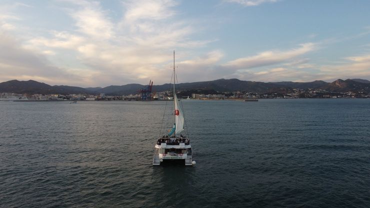 Velero en aguas tranquilas con una ciudad y colinas al fondo al atardecer.