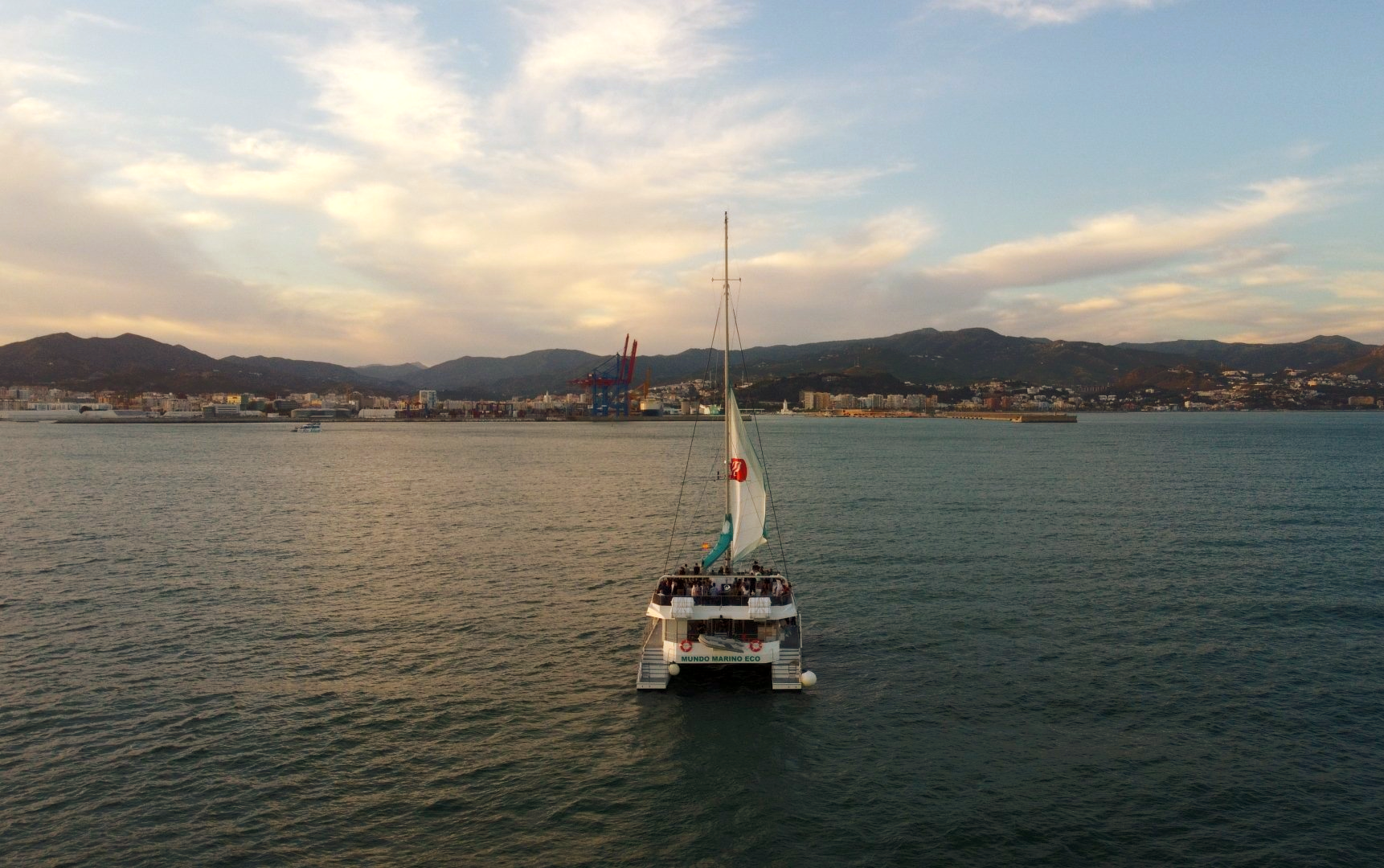 Un pequeño velero en aguas tranquilas con una ciudad y colinas al fondo al atardecer.