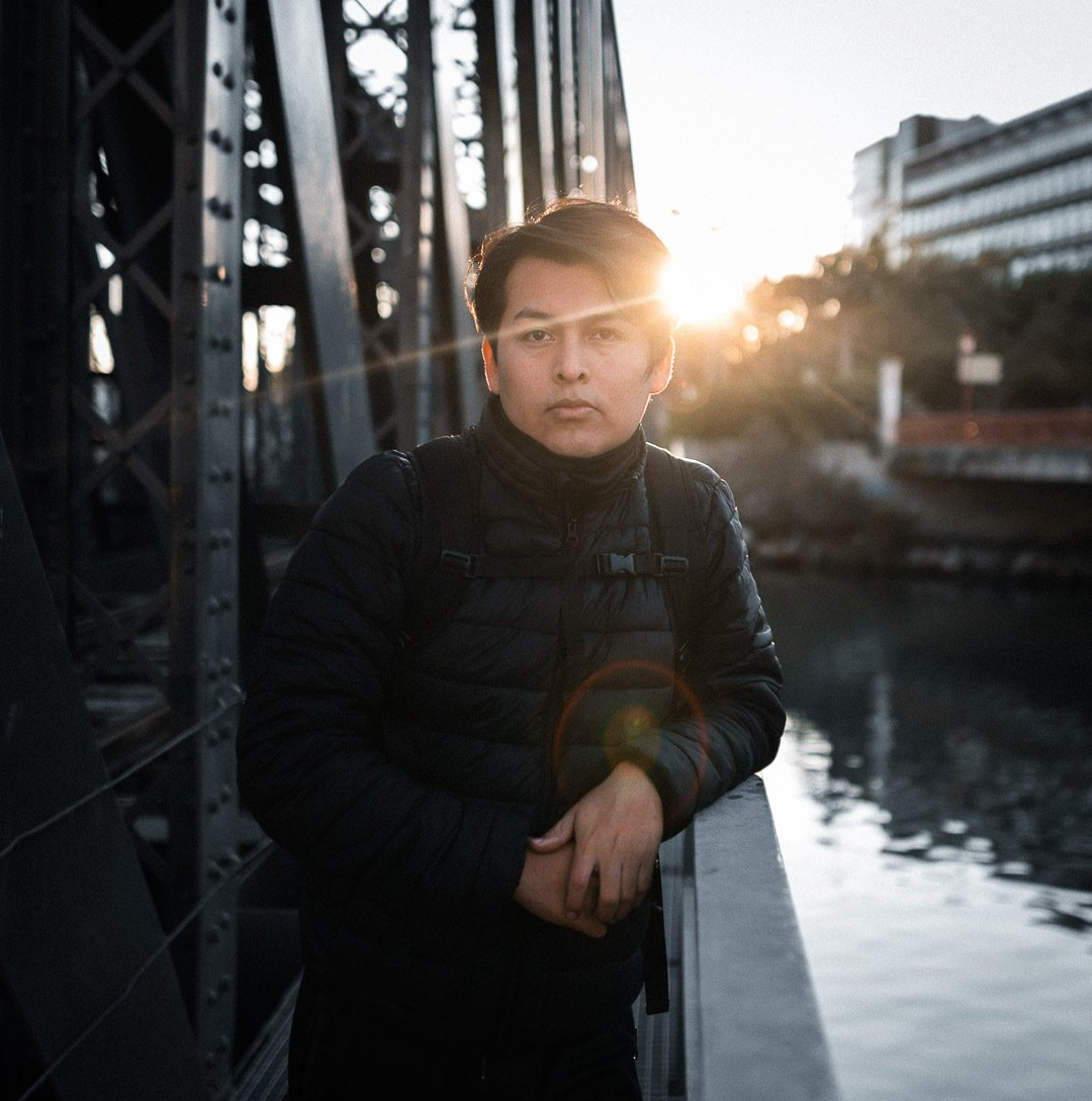 Una persona con un abrigo negro se encuentra de pie sobre un puente al atardecer, junto a un río, con edificios de la ciudad al fondo.