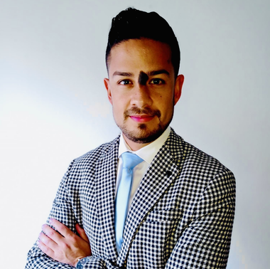 Retrato de una persona con traje a cuadros y corbata azul claro, con los brazos cruzados sobre un fondo blanco.