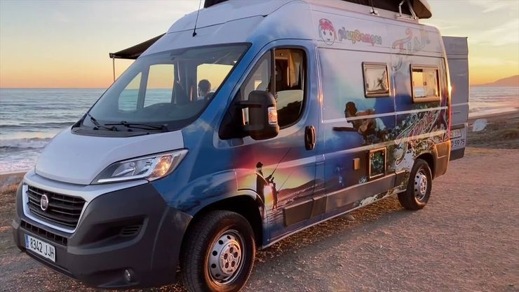 Una autocaravana azul aparcada en una playa al atardecer junto al agua.