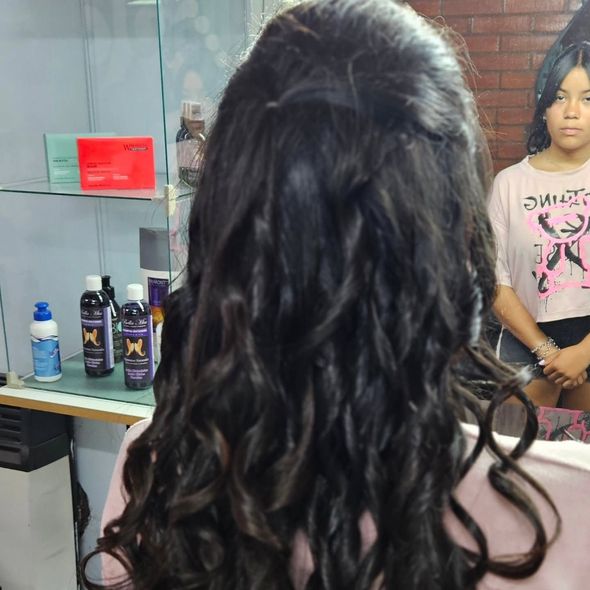Mujer con cabello oscuro, largo y ondulado peinado en un salón, frente a un espejo con otra mujer.