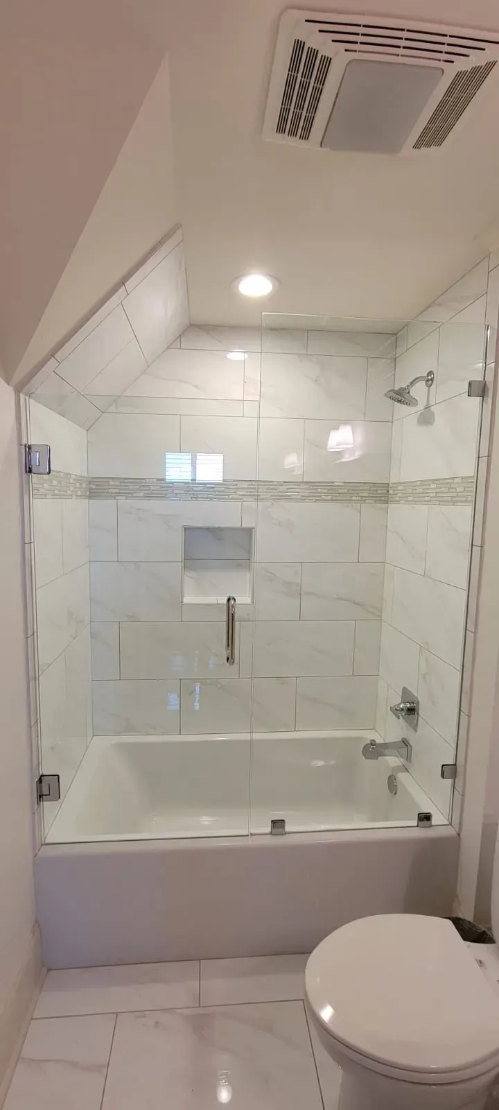 A bathroom with a white bathtub and glass shower door, white tile and a toilet.