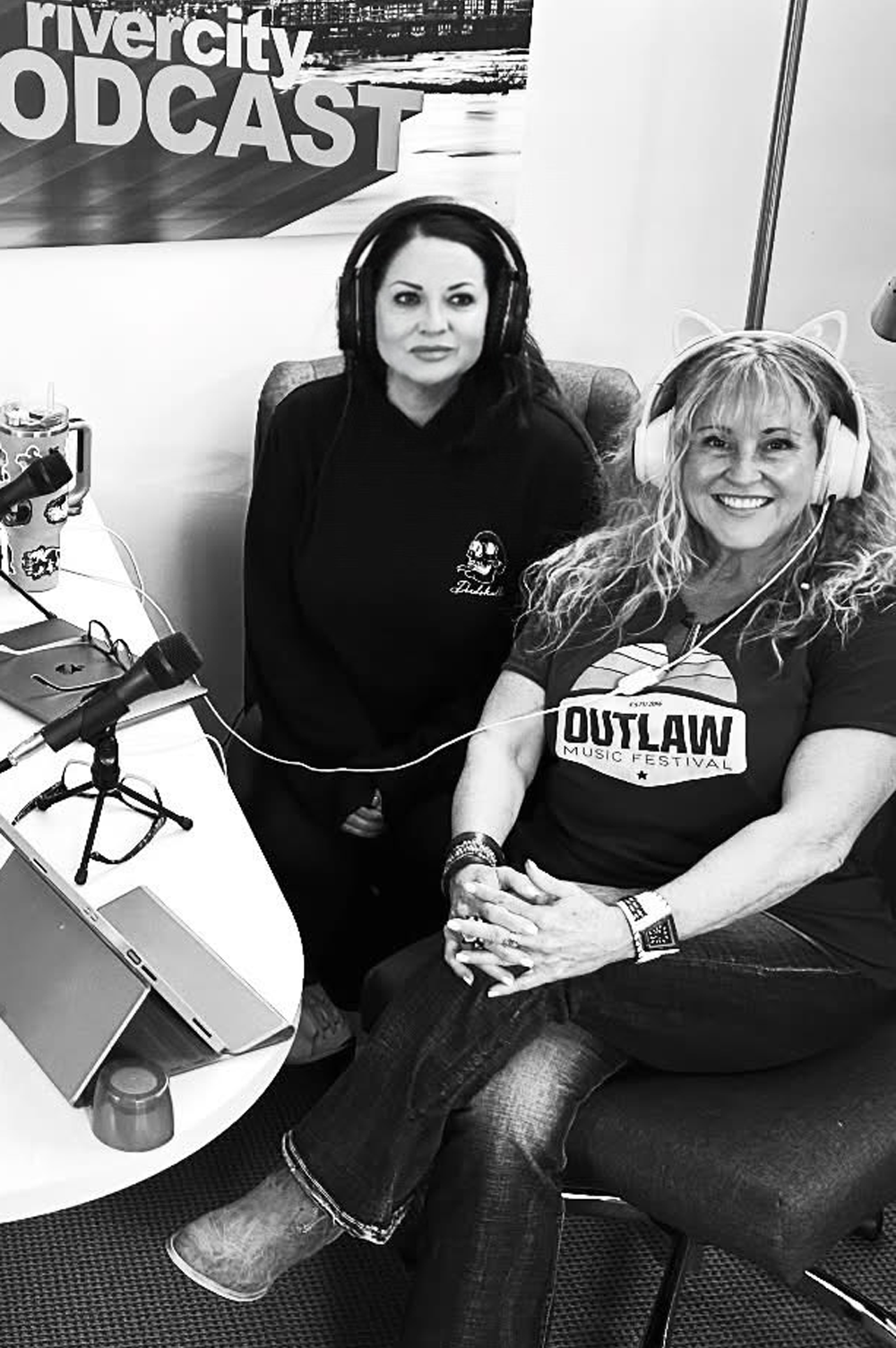 Two women at a podcast studio, one seated, wearing headphones, the other standing behind her. 