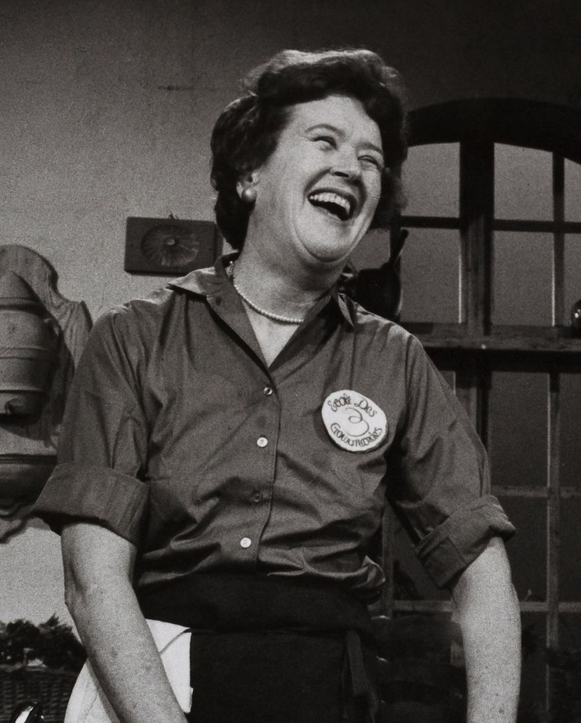 Julia Child laughing heartily in a kitchen, wearing a chef's uniform and apron.