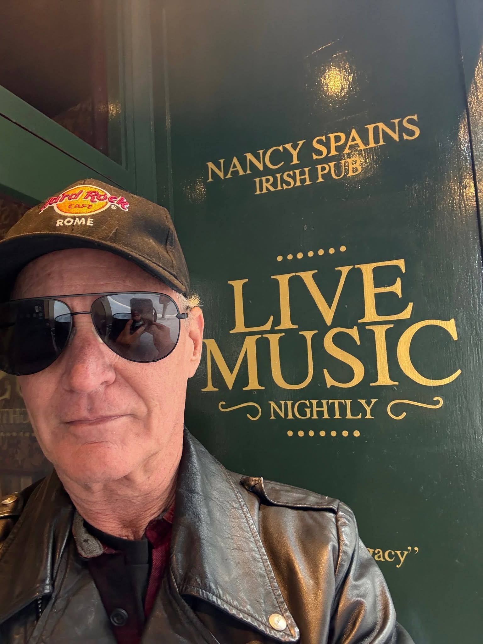 Man in sunglasses and cap posing by a Nancy Spungen live music sign