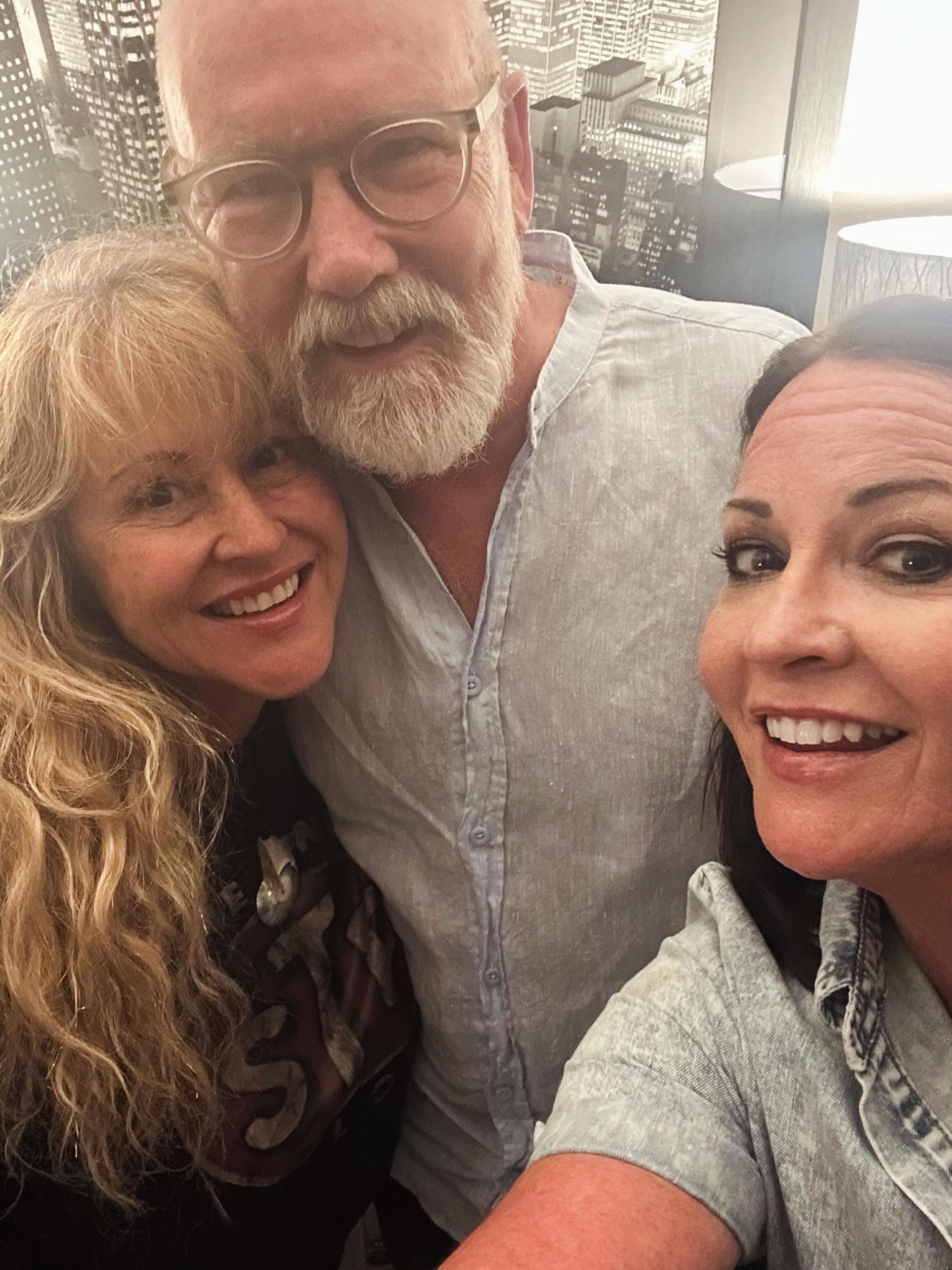 Three smiling adults posing closely together indoors, one taking the selfie.