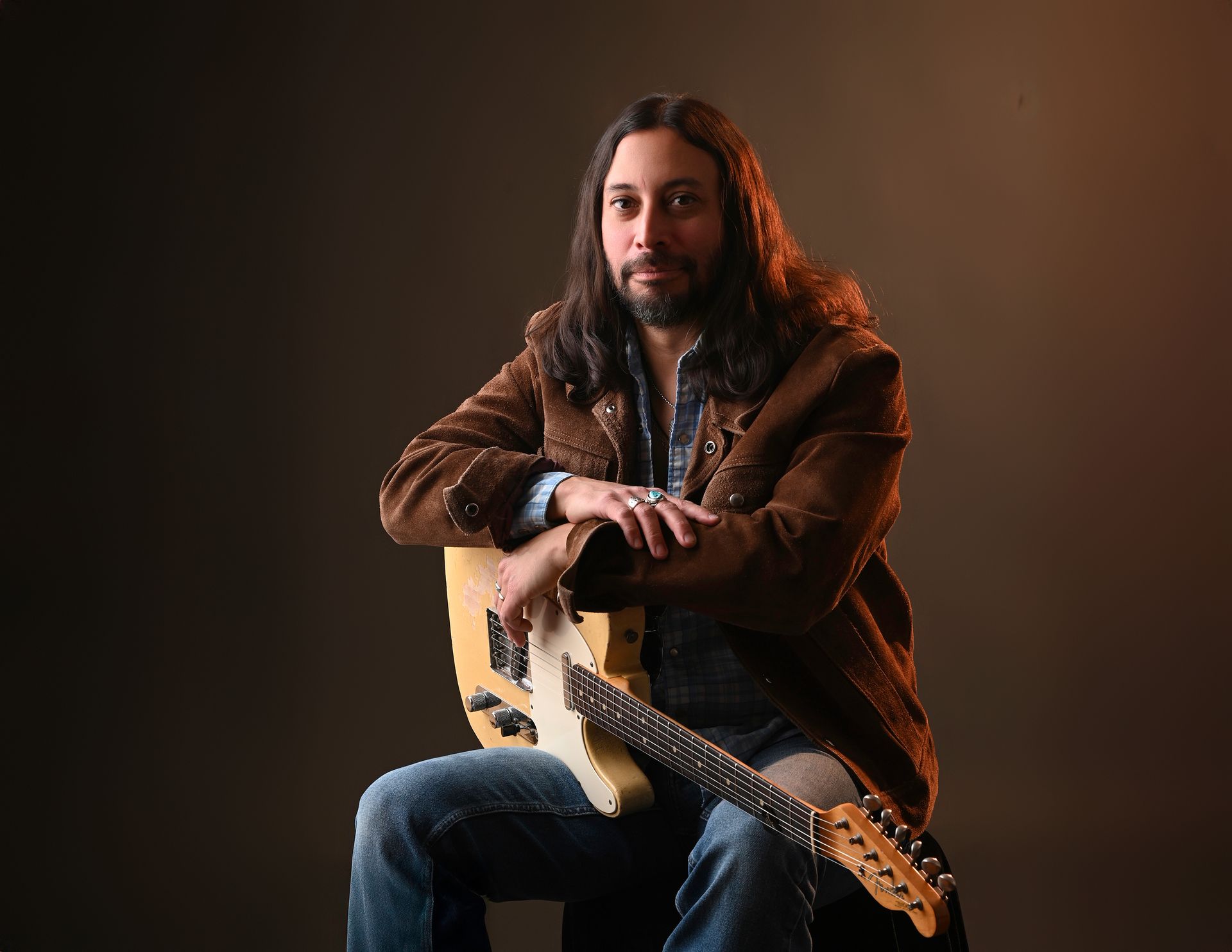 Man seated with electric guitar in a dark studio, wearing a brown jacket and jeans