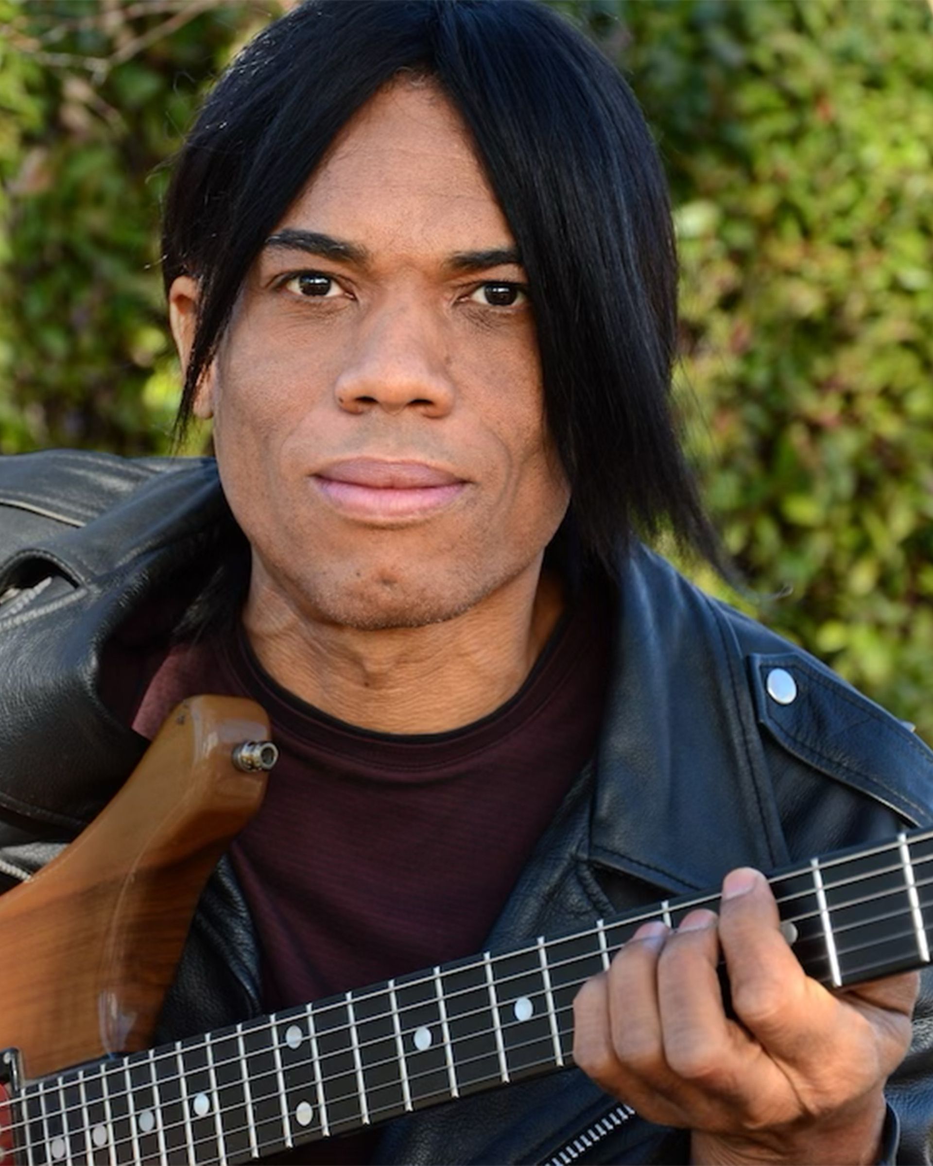 Person with black hair holding a guitar outdoors against green foliage.