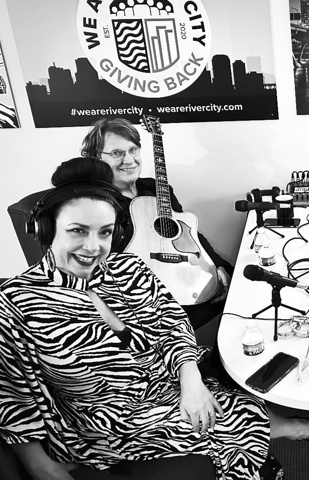 Four people at a table with microphones, recording a podcast. Studio setting, posters on the wall, guitar.